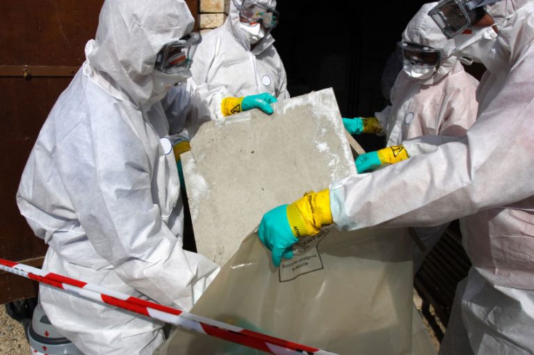 Group of people in asbestos hooded overalls putting asbestos in a bin bag behind red and white tape.